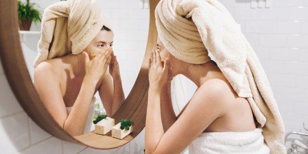 Young happy woman in towel applying organic face scrub and looking at round mirror in stylish bathroom. Girl making facial massage, peeling and cleaning skin on face. Skin Care and Hygiene